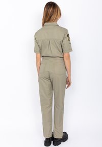 Monosuit caqui con mangas cortas, detalle de pliegue en la parte posterior y dos bolsillos traseros. Combinado con botas negras gruesas.