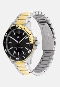 Herrklocka med svart urtavla, band av rostfritt stål i silver och guld, svart bezel och vita markörer; har varumärket "HILFIGER".