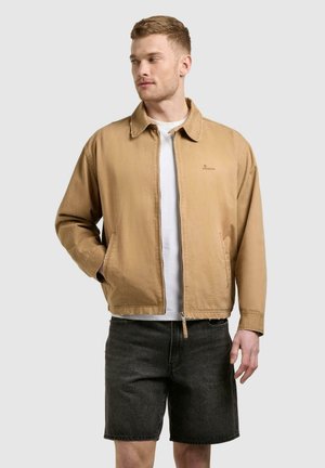 Homme portant une veste zippée beige sur un t-shirt blanc et un short en jean noir, debout avec une main dans la poche, regardant sur le côté.