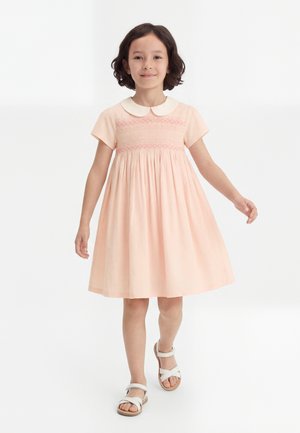 Jong meisje met donker haar, gekleed in een lichtroze jurk met een witte kraag en witte sandalen, lopend naar voren op een witte achtergrond.