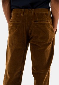 Bruine corduroy broek met een gestructureerd oppervlak, voorzien van twee achterzakken en een merklabel nabij de tailleband.