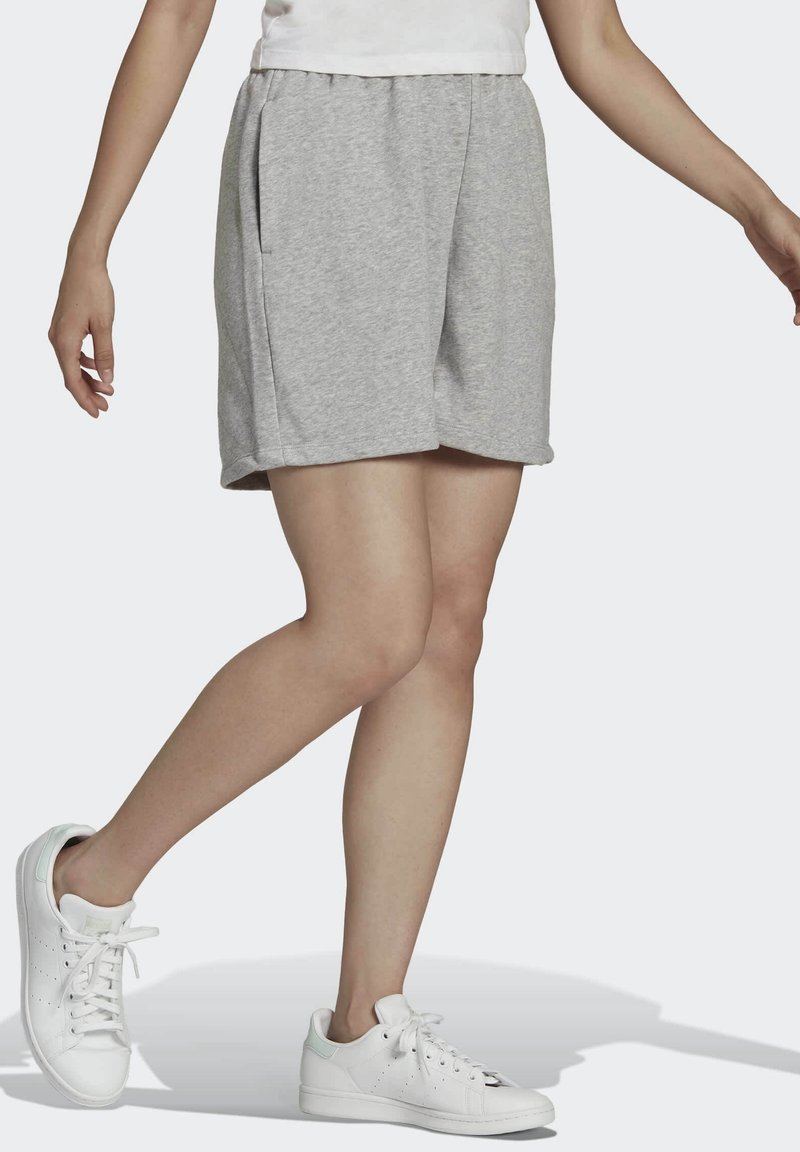 Osoba v světlých šedých šortkách a bílých teniskách, stojící s jednou nohou přehozenou přes druhou na jednobarevném pozadí.