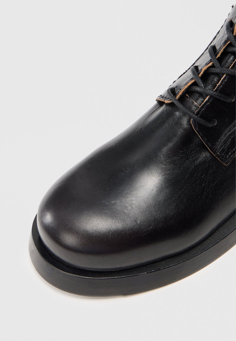 Black leather boot with a smooth finish, rounded toe, contrasting stitching, and a black rubber sole. Laces extend through eyelets on the side.