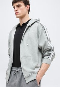 Ung mann iført en lys grå zip-up hettegenser med hvite og svarte striper på ermene, svart polo-skjorte og grå joggebukser med snøring.