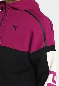 Sweatshirt à capuche en magenta vif et noir, avec un logo sur la poitrine, des manches blanches contrastantes avec un texte audacieux, et un tissu texturé.