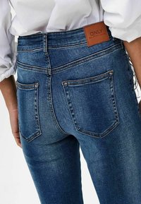 Gros plan sur une personne portant un jean denim bleu avec des poches arrière et un patch en cuir marron portant l'inscription « ONLY », associé à une chemise blanche.