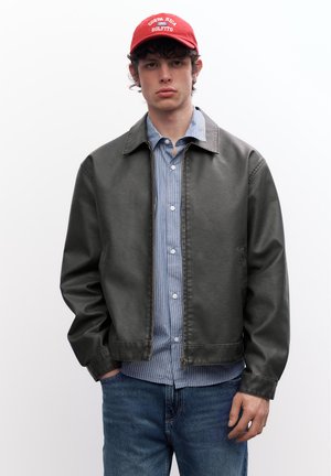 Tânăr purtând o șapcă roșie „Costa Rica Golfito”, geacă gri închis cu fermoar, cămașă albastru în dungi cu nasturi și blugi albaștri, cu o mână în buzunar.