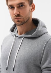 Felpa grigia realizzata in tessuto morbido, con tasca frontale, cappuccio con corde e accenti bianchi, e un design a vestibilità rilassata.