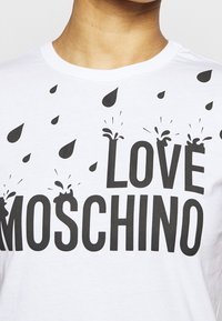 Bílý tričko s černým nápisem "Love Moschino" a grafikou kapek vody a stříkanců kolem písmen na oblasti hrudi.