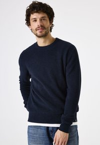 Navyblauwe gebreide trui met een gestructureerd oppervlak en ronde halslijn, gecombineerd met lichtblauwe jeans. Voorzien van geribde mouwen en onderkant.
