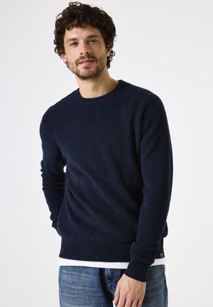 Navyblauwe gebreide trui met een gestructureerd oppervlak en ronde halslijn, gecombineerd met lichtblauwe jeans. Voorzien van geribde mouwen en onderkant.