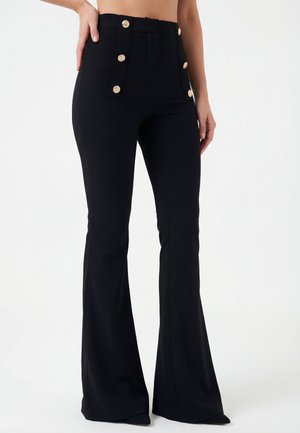 Czarne, rozszerzane spodnie z wysokim stanem i złotymi guzikami z przodu, noszone przez osobę z jedną ręką opartą na biodrze.