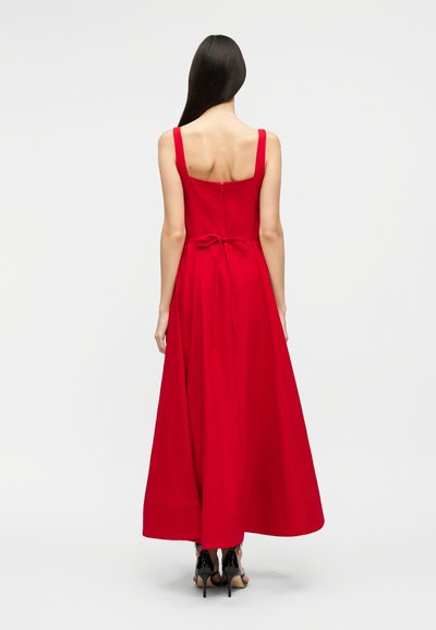 Vrouw met lang donker haar, gekleed in een mouwloze rode maxi-jurk met een gestrikt strikje in de taille, die met haar rug naar de camera staat tegen een effen witte achtergrond.