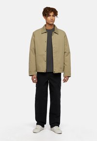 Khaki jacket with a snap button closure and green collar, worn over a dark shirt. Paired with black cargo pants and white sneakers.
