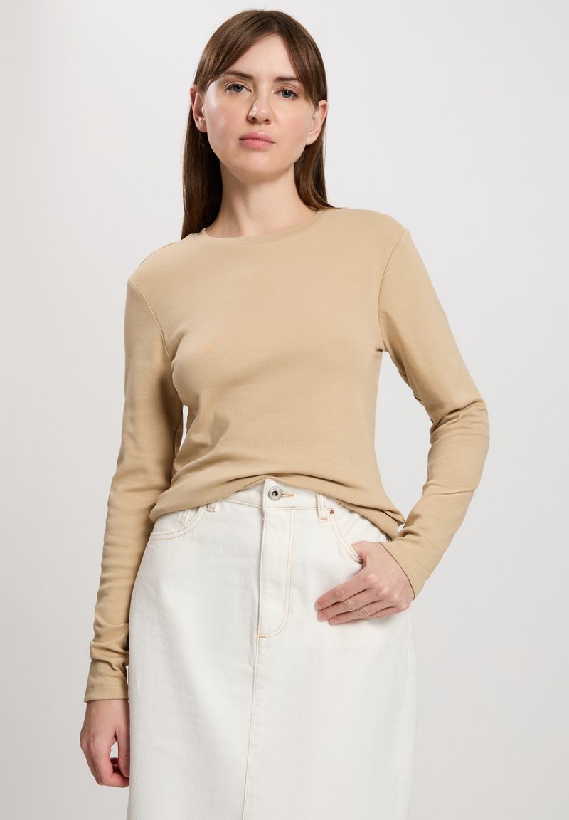 Beige Langarmshirt mit Rundhalsausschnitt, kombiniert mit einem weißen Denimrock mit einer Knopföffnung vorne und seitlichen Taschen.