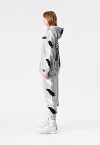 Persona de perfil vistiendo sudadera con capucha gris y pantalones con diseños de plumas en blanco y negro, combinados con botas blancas con cordones.