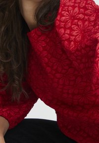 Blouse à motifs floraux rouges avec un tissu texturé et transparent. Présente des manches larges et une coupe décontractée, mettant en valeur des motifs de fleurs délicats.