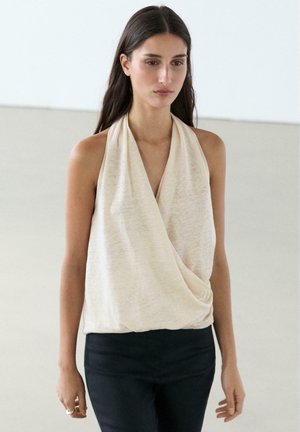 Donna con lunghi capelli scuri che indossa un top avvolgente beige senza maniche e pantaloni neri, in piedi contro uno sfondo chiaro semplice.