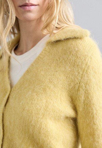 Κίτρινο μάλλινο πουλόβερ με μαλακή και φουντωτή υφή, διαθέτοντας γιακά και σχέδιο με V λαιμόκοψη, τοποθετημένο πάνω από ένα ανοιχτόχρωμο τοπ.