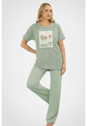 Femme portant un ensemble de pyjama vert clair avec un imprimé de palmier sur la chemise et de petits motifs de palmier sur le pantalon, debout avec une jambe pliée.