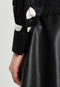 Blouse noire à texture avec des accents blancs à motifs, montrant un gros plan sur la manche et une jupe en cuir noir lisse. Détail des boutons visible.