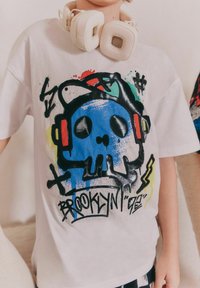 Vit bomulls t-shirt med en blå skallegrafik, färgglada stänk och texten "BROOKLYN '92." Hörlurar hänger runt nacken.