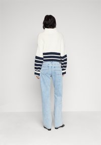 Persona vistiendo un suéter de rayas blanco y azul marino y unos vaqueros de tiro alto azules claros, de pie frente a una pared blanca lisa.