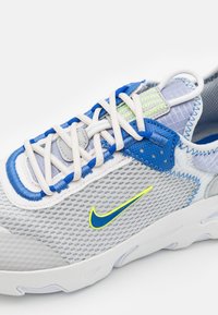 Šedé síťované tenisky s modrými a bílými akcenty, s výrazným zeleným logem Nike, textura povrchu a složitý systém šněrování.