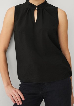 Blouse noire sans manches avec un petit col volanté et un décolleté en trou de serrure fermé par un seul bouton, portée avec un pantalon foncé.