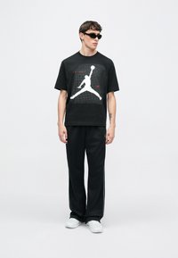 Μαύρο μπλουζάκι με λευκό λογότυπο Jordan, συνδυασμένο με μαύρο παντελόνι φόρμας και λευκά αθλητικά παπούτσια. Γυαλιά ηλίου ολοκληρώνουν το casual look.