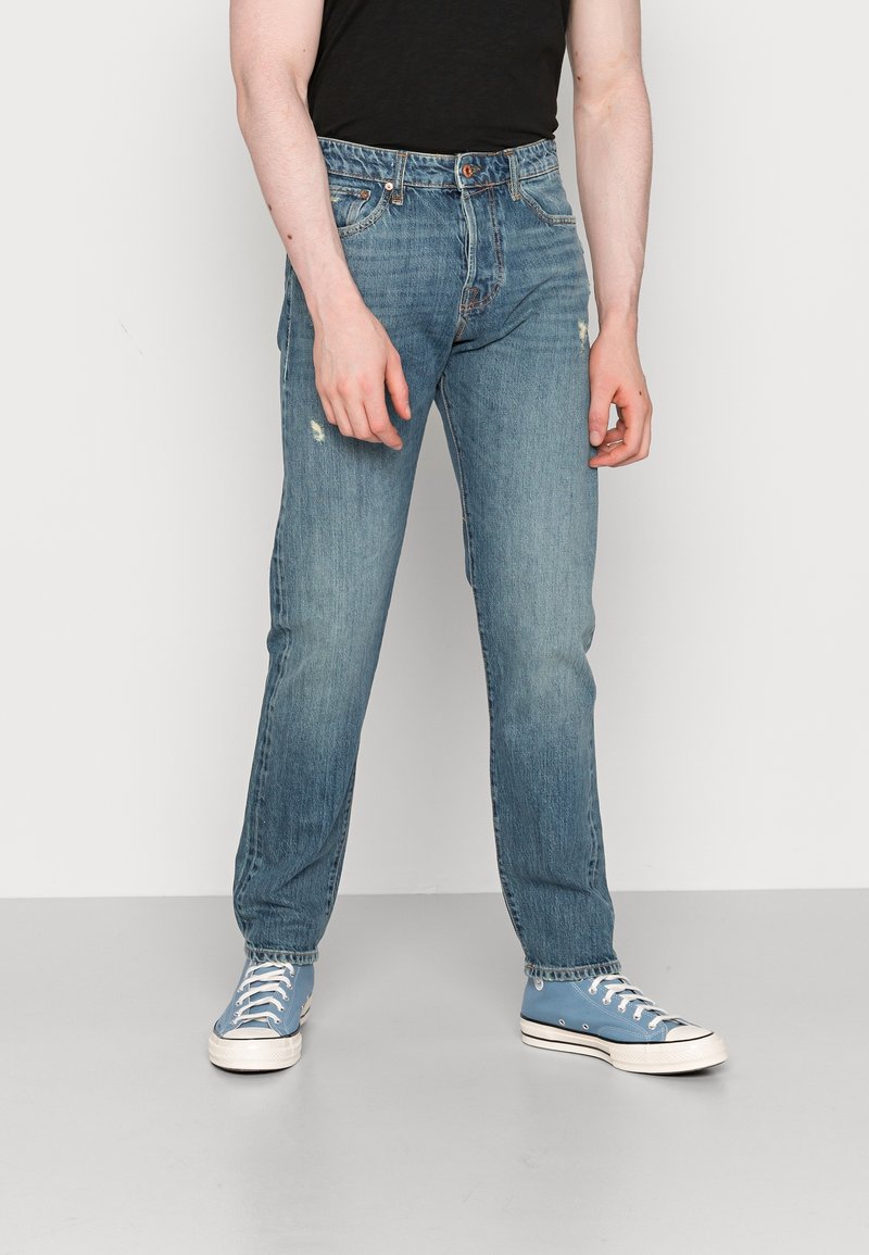 Jeans de denim azul claro con un corte recto, que presentan un ligero desgaste y un diseño tradicional de cinco bolsillos. Llevados con zapatillas azules.
