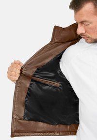 Giacca in pelle marrone con una texture liscia, dotata di un interno in satin nero e una tasca con zip sul lato sinistro.