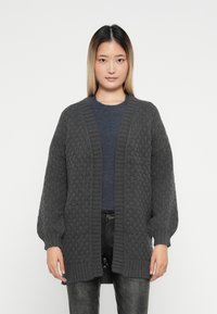 Grå strikket cardigan med et teksturert diamantmønster, åpen foran, ribbestrikkede mansjetter og hem, båret over en mørk topp.