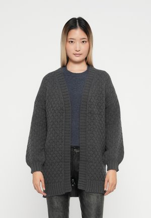 Šedý pletený cardigan s texturovaným diamantovým vzorem, otevřený vpředu, s žebrovanými manžetami a lemem, nošený přes tmavý top.