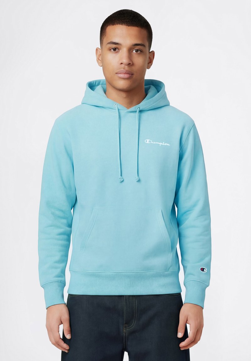Jonge man die een lichtblauwe Champion-hoodie draagt met een voorzak en logo op de borst en linker pols, staand tegen een effen witte achtergrond.