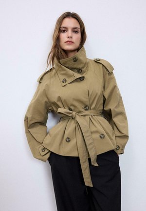 Ženska, oblečena v bež plašč s pasom, velikimi gumbi in visokim ovratnikom, stoji pred enobarvnim belim ozadjem.