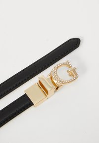 Zwarte lederen riem met een glanzende gouden gesp in de vorm van de letter 'G', versierd met kleine rhinestones rond de randen.