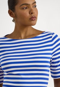 Woman with smooth bun wearing round gold earrings and a blue and white horizontal striped top with a wide neckline.