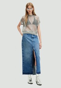 Šedý síťovaný top s krátkými rukávy, spárovaný s maxi sukní z modrého denimu s vysokým pasem, která má přední rozparek. Model má na sobě lesklé stříbrné boty.