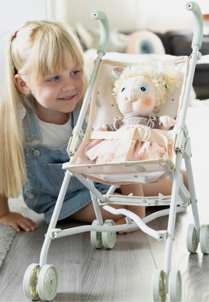 Jeune fille aux cheveux blonds sourit en s'agenouillant à côté d'une poussette jouet tenant une poupée habillée de vêtements roses et beiges.