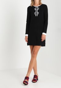 Žena v černých mini šatech s dlouhým rukávem a bílými lemy, v červených sandálech na podpatku se vzorem, stojí před jednobarevným bílým pozadím.