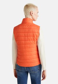 Gilet imbottito arancione con colletto alto, motivo a righe orizzontali e materiale leggero, abbinato a una maglietta bianca a maniche lunghe e jeans.