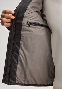 Eine Hand hält eine offene, dunkelbraune Lederjacke, die ein hellgraues Innenfutter mit einer Reißverschlusstasche auf der rechten Seite zeigt.