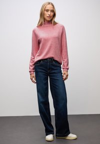 Rosa gestrickter Pullover mit hochgeschlossenem Kragen, kombiniert mit dunkelblauen weiten Jeans. Der Stoff wirkt weich, mit gerippten Bündchen und einer lässigen Passform.