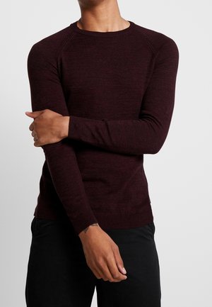 Homme portant un pull ajusté à manches longues col rond de couleur bordeaux foncé et un pantalon noir, avec un bras croisé sur l'autre.