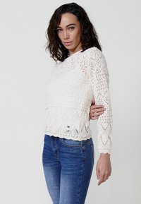 Suéter blanco de punto con un patrón de encaje, mangas largas y detalles texturizados; combinado con jeans azules. Presenta sutiles fruncidos en el dobladillo.