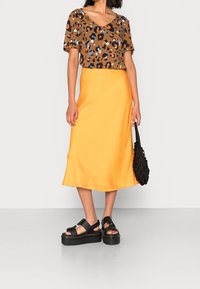 Haut à manches courtes en imprimé léopard marron, jupe midi en satin jaune, sandales noires à semelle épaisse et sac à main tissé noir. Textures lisses et couleurs vives.