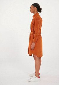 Robe chemise orange rouille avec des manches longues, une taille ceinturée, des poches latérales et un ourlet arrondi. Associée à des baskets blanches avec des accents rouges.