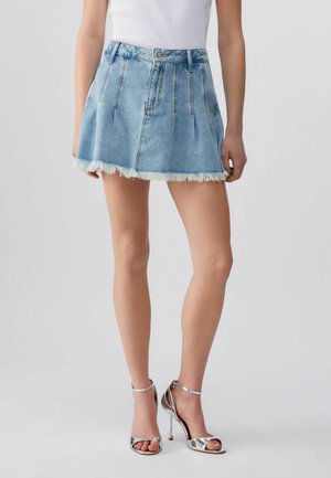 Donna che indossa una minigonna di jeans azzurro chiaro sfrangiata, abbinata a sandali argento con tacco alto e cinturino alla caviglia, in piedi davanti a uno sfondo neutro.