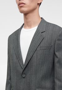 Jeune homme portant un blazer gris à motif chevron sur un t-shirt blanc à col rond, visible des épaules au menton.